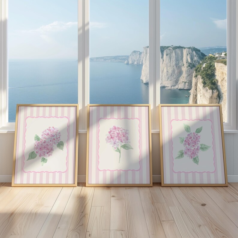 May include: Three framed floral art prints featuring watercolor hydrangeas. Each print has a pink and white striped border and a gold frame. The background shows a scenic view of the ocean and cliffs.