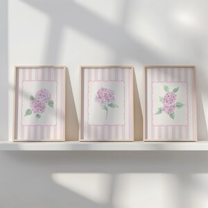 May include: Three framed floral art prints featuring watercolor paintings of pink hydrangeas. The prints have a striped background with a pink border and are displayed on a white shelf.