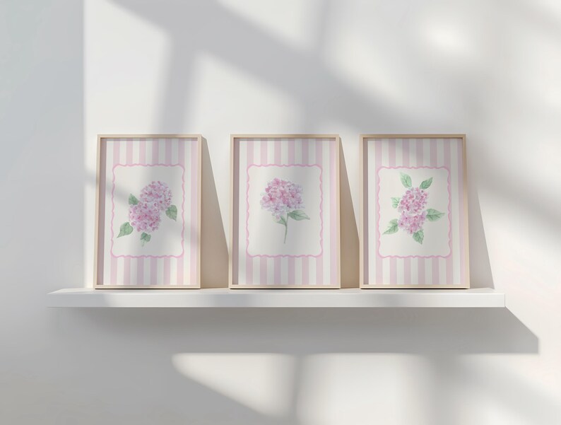May include: Three framed floral art prints featuring watercolor paintings of pink hydrangeas. The prints have a striped background with a pink border and are displayed on a white shelf.