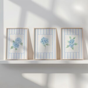 May include: Three framed art prints featuring watercolor paintings of blue hydrangeas. Each print has a light blue and white striped background with a white scalloped border around the floral image. The frames are a light wood color.