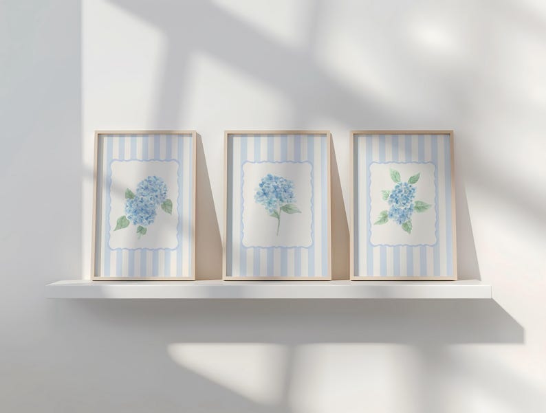 May include: Three framed art prints featuring watercolor paintings of blue hydrangeas. Each print has a light blue and white striped background with a white scalloped border around the floral image. The frames are a light wood color.