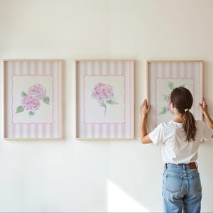 May include: Three framed floral art prints featuring watercolor hydrangea blooms in shades of pink and green. The prints have a striped background and are displayed on a white wall. The frames are light wood.