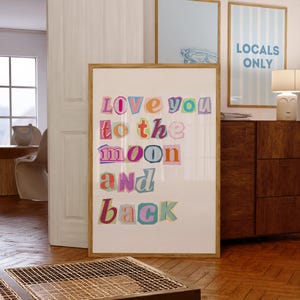 以下が含まれることがあります： カラフルな切り抜き文字で「Love you to the moon and back」と書かれた額入りのアートプリント。他の額入りのアートや木製家具のある部屋に飾られています。