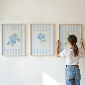 May include: Three framed floral art prints featuring blue hydrangea blooms. The prints have a light blue and white striped background with a scalloped border. The frames are a light wood color.