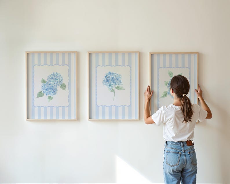 May include: Three framed floral art prints featuring blue hydrangea blooms. The prints have a light blue and white striped background with a scalloped border. The frames are a light wood color.