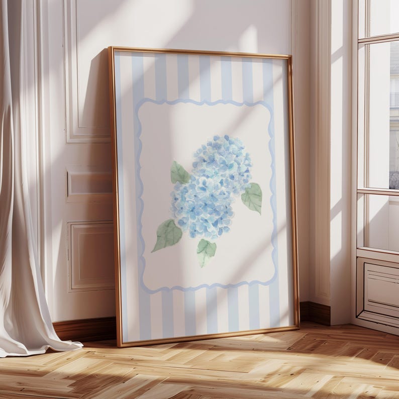 May include: A framed art print featuring a watercolor painting of blue hydrangea flowers with green leaves. The artwork is set against a white background with a light blue striped border and a gold frame. The print is leaning against a wall.