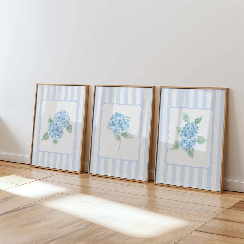 May include: Three framed art prints featuring watercolor paintings of blue hydrangea flowers. Each print has a white background with a blue floral design, set against a light blue and white striped background. The frames are a warm, natural wood tone.