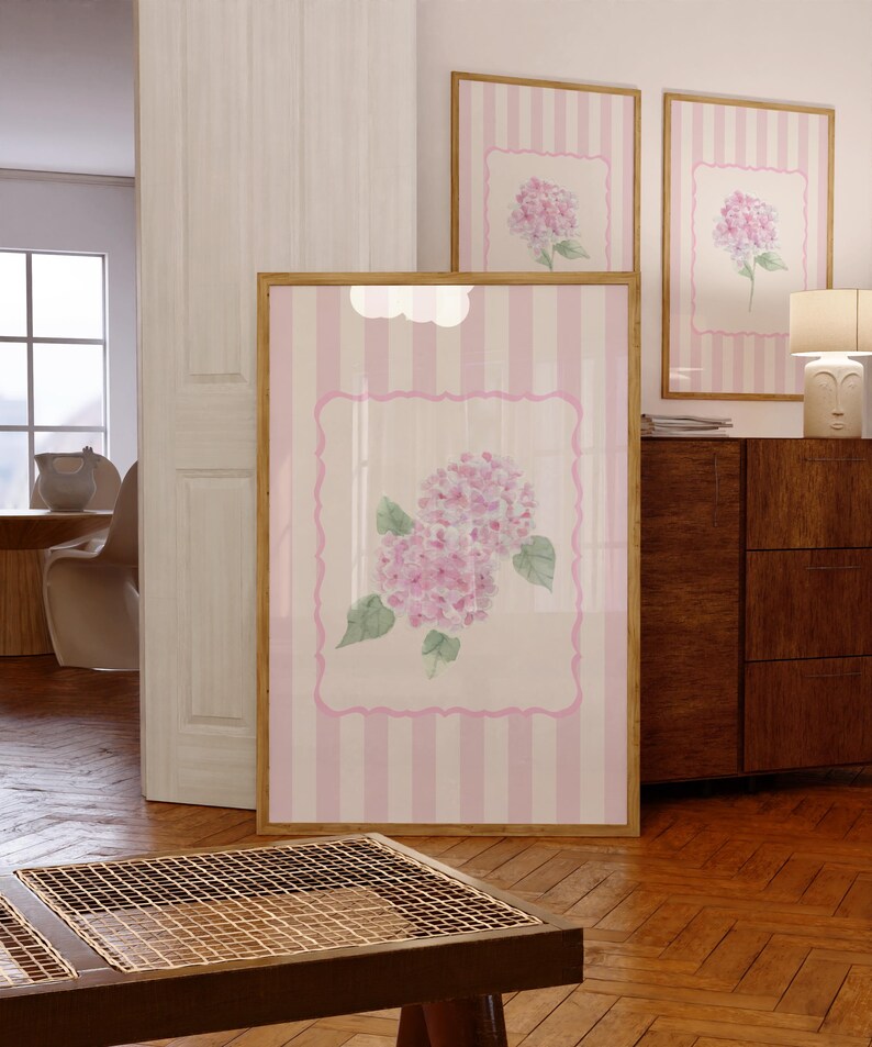 May include: Three framed floral art prints. The prints feature pink hydrangea flowers on a cream background with a pink striped border. The frames are a light brown color. The art is displayed in a room with hardwood floors.