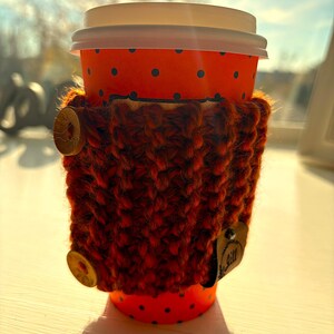 Op de afbeelding: Een rode en witte papieren koffiebeker met stippen en een bruin gebreide bekerhoes. De bekerhoes heeft twee bruine knopen en een klein bruin label met het woord "Gill" erop.