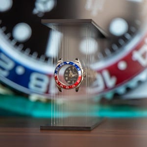 May include: A stainless steel watch bezel with a red and blue rotating ring, displayed on a clear acrylic stand. The watch face is visible through the bezel, with a black dial and luminous markers. The background features a blurred watch face.