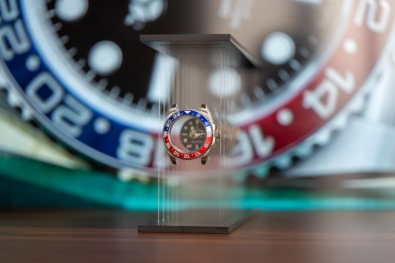 May include: A stainless steel watch bezel with a red and blue rotating ring, displayed on a clear acrylic stand. The watch face is visible through the bezel, with a black dial and luminous markers. The background features a blurred watch face.