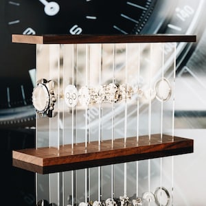 May include: A watch parts display stand featuring a series of clear vertical acrylic panels held between two horizontal dark wood planks. The transparent panels are arranged to hold various metallic watch components in a linear sequence.