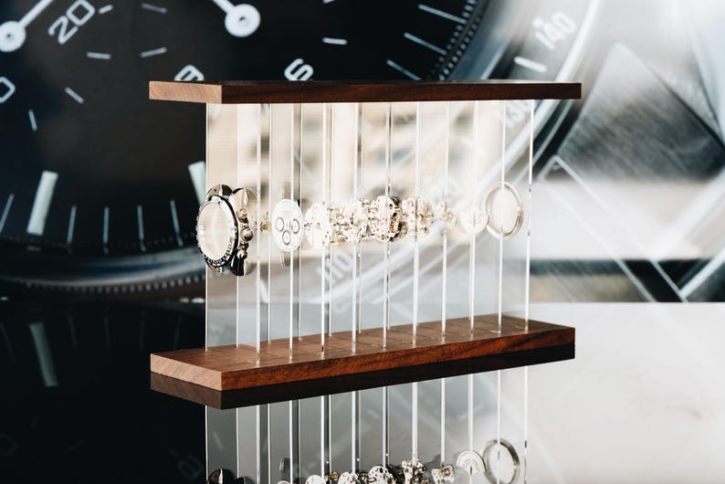 May include: A watch parts display stand featuring a series of clear vertical acrylic panels held between two horizontal dark wood planks. The transparent panels are arranged to hold various metallic watch components in a linear sequence.