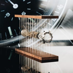 May include: A disassembled silver wristwatch with its internal components, including the case, dial, and gears, suspended horizontally between two parallel dark wood rectangular blocks by thin, vertical, transparent nylon wires.