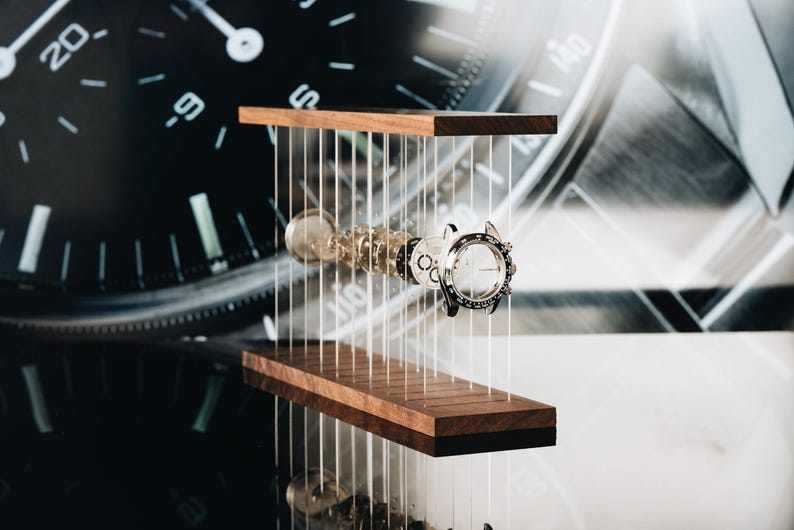 May include: A disassembled silver wristwatch with its internal components, including the case, dial, and gears, suspended horizontally between two parallel dark wood rectangular blocks by thin, vertical, transparent nylon wires.