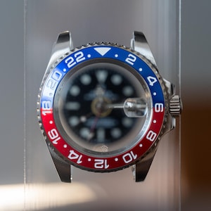May include: A stainless steel wristwatch with a black dial and a bi-directional rotating bezel in red and blue. The watch has white numerals and markers, and a crown on the right side.