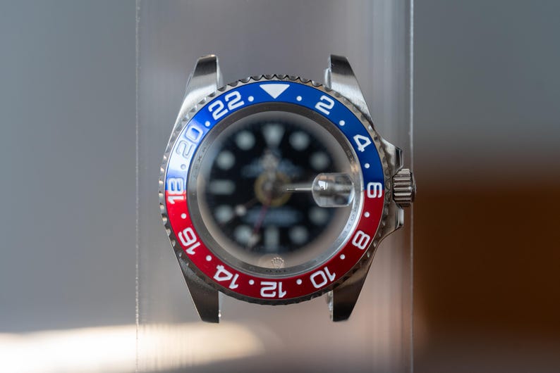 May include: A stainless steel wristwatch with a black dial and a bi-directional rotating bezel in red and blue. The watch has white numerals and markers, and a crown on the right side.