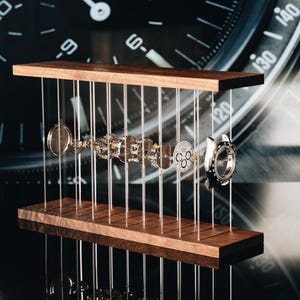 May include: A disassembled mechanical watch movement displayed in an exploded view between two horizontal wooden planks connected by thin vertical wires. The metallic watch components are suspended in a row, showcasing the internal gears and casing.