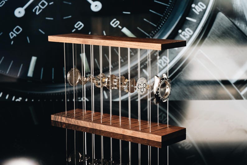 May include: A disassembled mechanical watch movement displayed in an exploded view between two horizontal wooden planks connected by thin vertical wires. The metallic watch components are suspended in a row, showcasing the internal gears and casing.