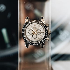 May include: A silver-tone chronograph wristwatch featuring a white dial with three sub-dials, a black tachymeter bezel with white markings, and a stainless steel case. The watch is mounted on a clear acrylic stand, displaying a classic circular design.