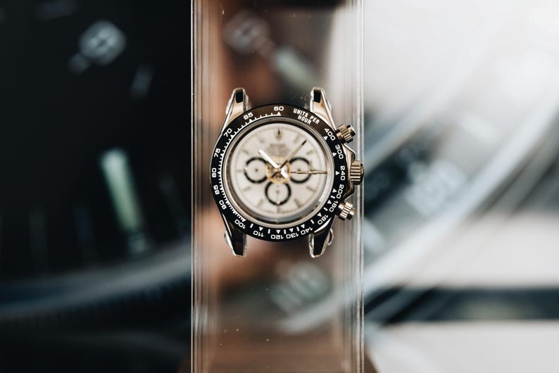 May include: A silver-tone chronograph wristwatch featuring a white dial with three sub-dials, a black tachymeter bezel with white markings, and a stainless steel case. The watch is mounted on a clear acrylic stand, displaying a classic circular design.