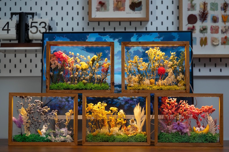May include: Five wooden framed floral arrangements. Each frame contains dried flowers and greenery, with a backdrop of a blue sky. The arrangements feature a variety of colours, including red, yellow, and purple, and are displayed on a wooden surface.