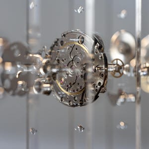 May include: Close-up of a watch mechanism with intricate gears and components. The silver-colored parts are arranged in a circular pattern, with a gold-colored central gear. The image has a shallow depth of field, with blurred elements in the foreground and background.