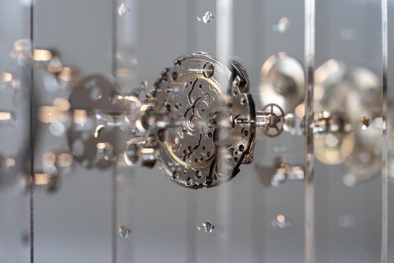 May include: Close-up of a watch mechanism with intricate gears and components. The silver-colored parts are arranged in a circular pattern, with a gold-colored central gear. The image has a shallow depth of field, with blurred elements in the foreground and background.