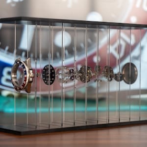 May include: A watch display stand showcasing the intricate internal components of a wristwatch. The stand is black, with clear vertical supports holding the watch parts in a suspended arrangement. The watch face has a blue and red bezel.