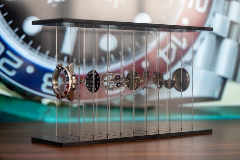 May include: A watch display stand showcasing the intricate internal components of a wristwatch. The stand is black, with clear vertical supports holding the watch parts in a suspended arrangement. The watch face has a blue and red bezel.