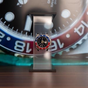 May include: A luxury wristwatch with a black dial, gold hands, and a red and blue bezel. The watch is displayed on a clear stand against a blurred background of a watch face with white numerals.