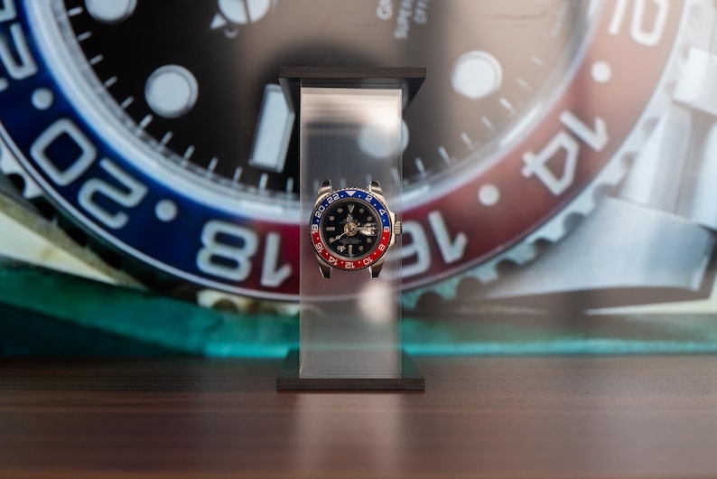 May include: A luxury wristwatch with a black dial, gold hands, and a red and blue bezel. The watch is displayed on a clear stand against a blurred background of a watch face with white numerals.