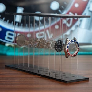 Może przedstawiać: Czarny stojak ekspozycyjny prezentujący zdemontowane elementy zegarka. Stojak posiada szereg przezroczystych, pionowych podpór trzymających części zegarka, w tym ramkę z czerwonymi i niebieskimi akcentami. W tle tarcza zegarka.