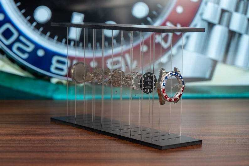 May include: A black display stand showcasing the disassembled components of a wristwatch. The stand features a series of clear vertical supports holding the watch parts, including the bezel with red and blue accents. The background shows a watch face.
