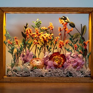 May include: A wooden framed display case featuring dried flowers. The arrangement includes orange, yellow, and pink flowers, with green foliage and white moss. The frame has a warm, natural wood tone, and is rectangular.