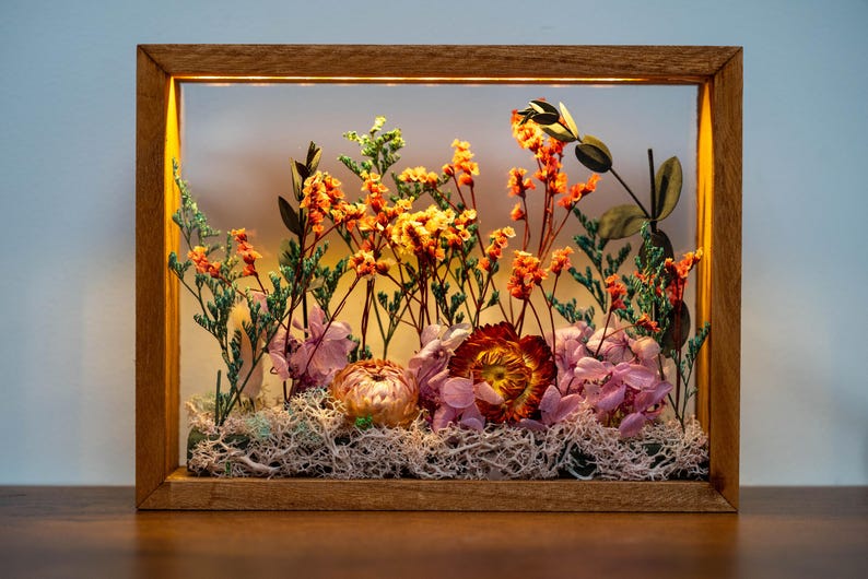 May include: A wooden framed display case featuring dried flowers. The arrangement includes orange, yellow, and pink flowers, with green foliage and white moss. The frame has a warm, natural wood tone, and is rectangular.