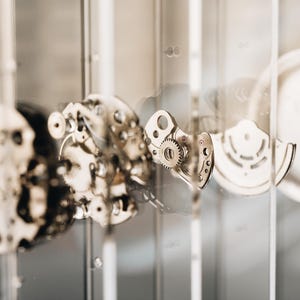 May include: A disassembled mechanical watch movement featuring various metallic gears, plates, and a circular rotor, all suspended between thin, transparent vertical glass panels that provide a clear view of the intricate internal components.