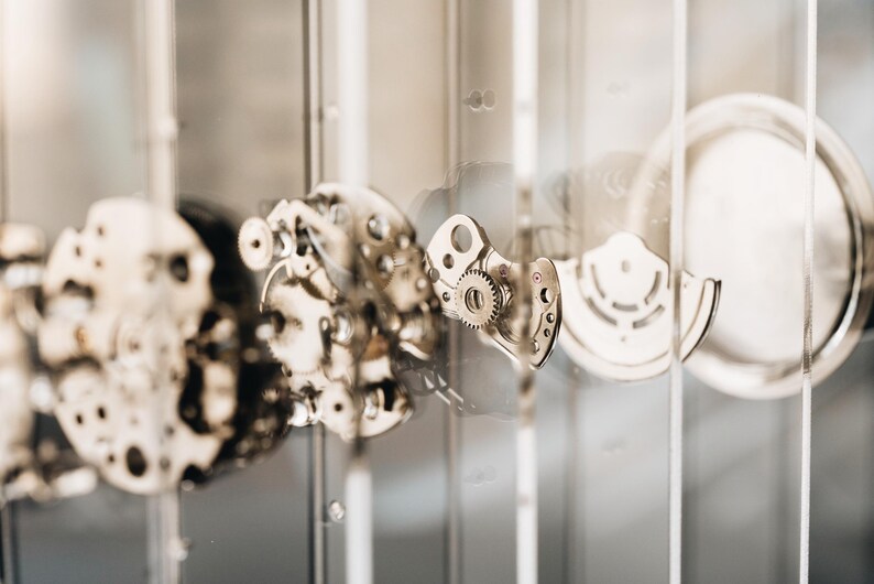 May include: A disassembled mechanical watch movement featuring various metallic gears, plates, and a circular rotor, all suspended between thin, transparent vertical glass panels that provide a clear view of the intricate internal components.