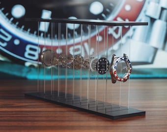 May include: An exploded view display of a wristwatch featuring a red and blue bezel, a black dial, and various internal mechanical components suspended between a black base and a top plate by thin, clear vertical wires in a linear arrangement.