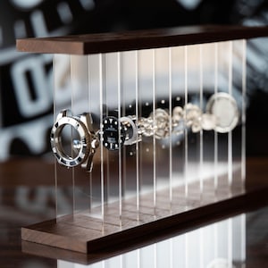 Peut inclure: Une montre mécanique démontée dont les composants sont suspendus en ligne entre deux bases horizontales en bois sombre. Les pièces internes, y compris le cadran avec le logo Rolex, sont maintenues par des panneaux en acrylique transparent.