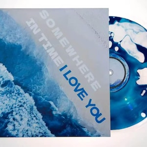 May include: A square album cover featuring an aerial view of blue ocean waves crashing onto a white sandy beach with the text SOMEWHERE IN TIME I LOVE YOU. Beside it is a circular vinyl record with a marbled blue and white liquid pattern.