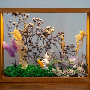 May include: A wooden framed display with a white rabbit figurine holding a carrot, set amongst dried flowers, green moss, and purple feathers. The frame is made of wood and has a warm tone.
