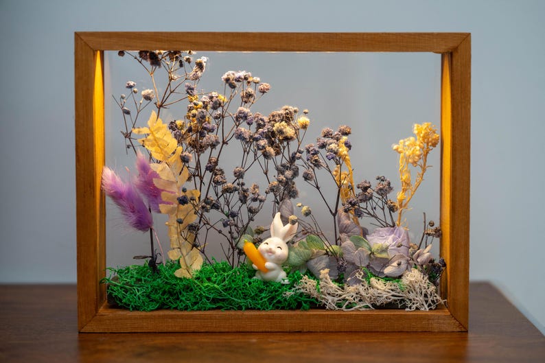 May include: A wooden framed display with a white rabbit figurine holding a carrot, set amongst dried flowers, green moss, and purple feathers. The frame is made of wood and has a warm tone.