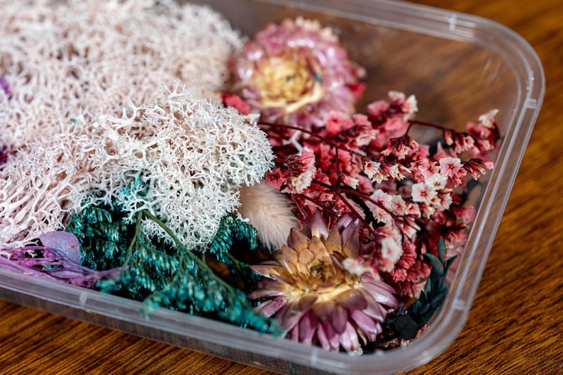 May include: A clear plastic container filled with dried flowers and foliage. The arrangement features white, teal, pink, and purple elements, creating a colourful and textured display. The flowers are likely for crafting or decorative purposes.