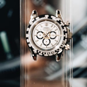 May include: A silver-tone chronograph wristwatch featuring a white dial with three black sub-dials, a black tachymeter bezel, and metallic hour markers. The watch case has two pushers on the right side and is displayed on a clear vertical stand.