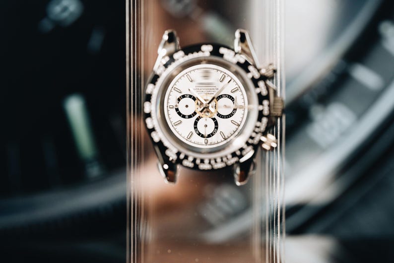 May include: A silver-tone chronograph wristwatch featuring a white dial with three black sub-dials, a black tachymeter bezel, and metallic hour markers. The watch case has two pushers on the right side and is displayed on a clear vertical stand.