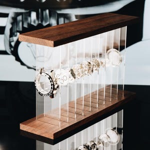 May include: A display stand featuring two horizontal dark wood planks connected by vertical clear acrylic panels. Suspended between the panels are disassembled watch components, including a circular watch case and various internal gear parts.