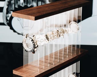 May include: A display stand featuring two horizontal dark wood planks connected by vertical clear acrylic panels. Suspended between the panels are disassembled watch components, including a circular watch case and various internal gear parts.