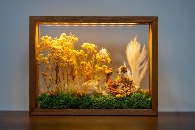 May include: A decorative framed floral display. The wooden frame surrounds a glass panel, illuminating dried yellow and white flowers, green moss, and a miniature violin. The arrangement is a natural, decorative piece.