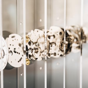 May include: A disassembled watch movement displayed behind vertical glass panels. The white watch face features three black sub-dials and numerical markers. Metallic gears, plates, and internal components are arranged in a linear sequence.
